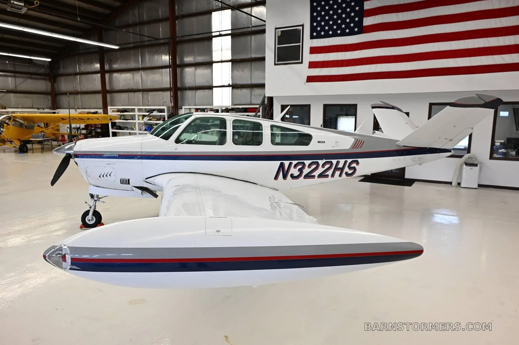 Side-rear view of a white single-engine aircraft with blue and red stripes, registration N322HS, in a hangar.