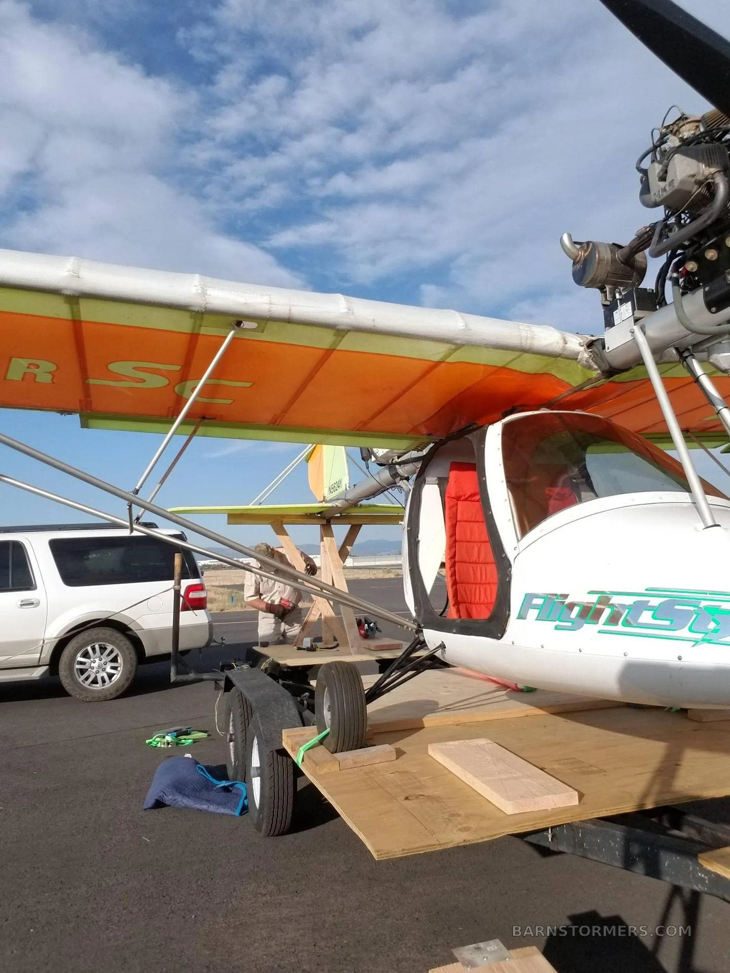 A white and orange ultralight aircraft with an open cockpit, parked on a tarmac next to a white SUV.