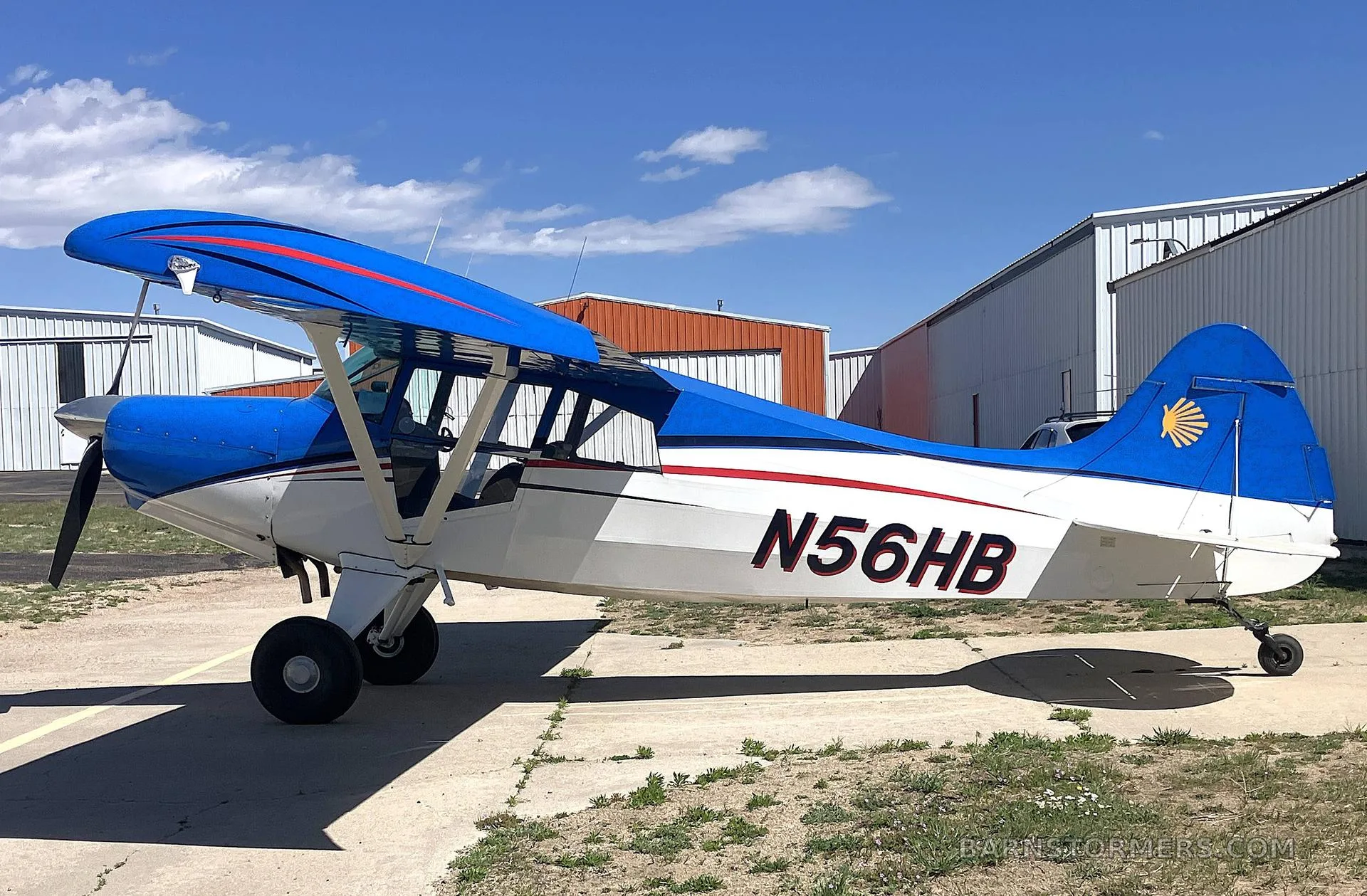 A blue and white high-wing aircraft with red stripes and tail logo, registration N56HB, parked on an asphalt apron with hangars in the background.