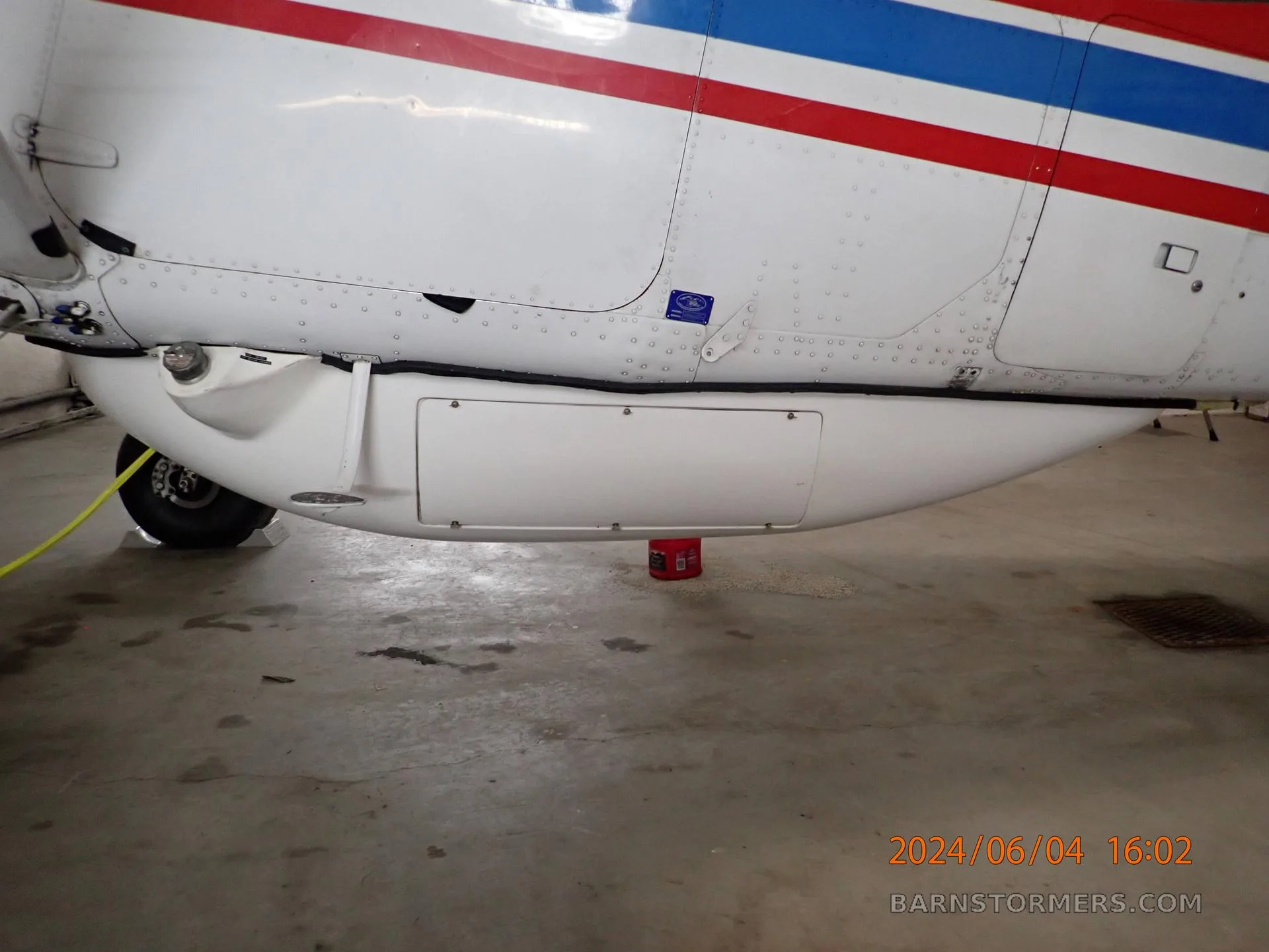 A close-up view of the white belly pod and landing gear of a red, white, and blue aircraft inside a hangar.