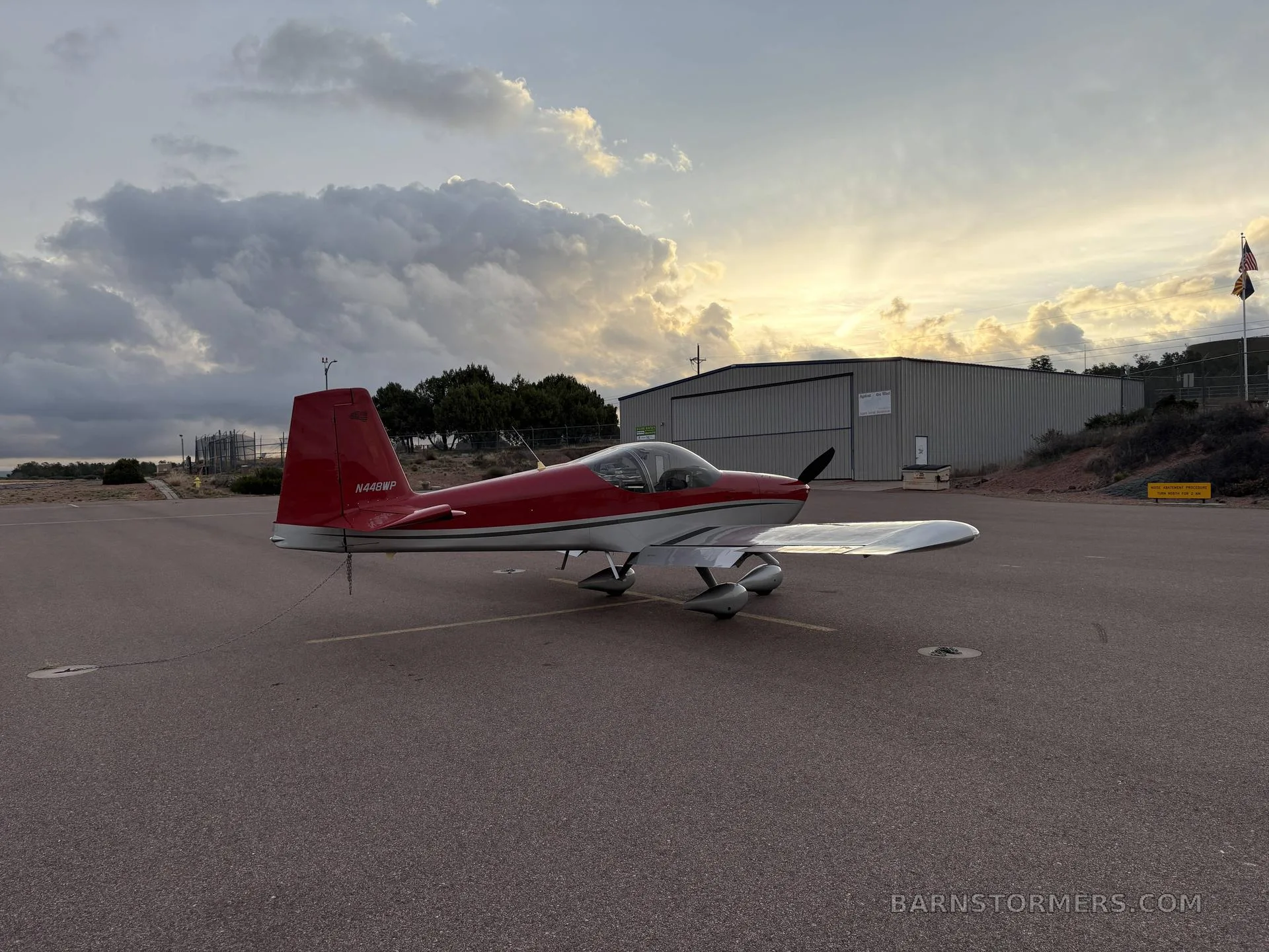 A red and silver single-engine aircraft, N448WP, tied down on an airfield with a hangar and sunset in the background.