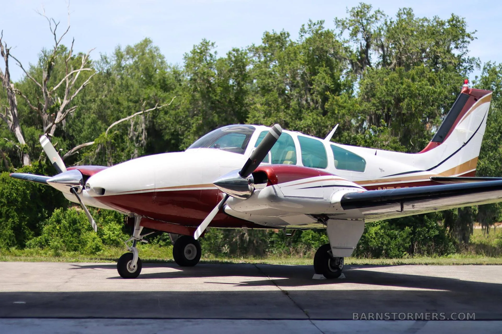 A white and maroon twin-engine propeller aircraft with gold stripes is parked on a tarmac with trees in the background.