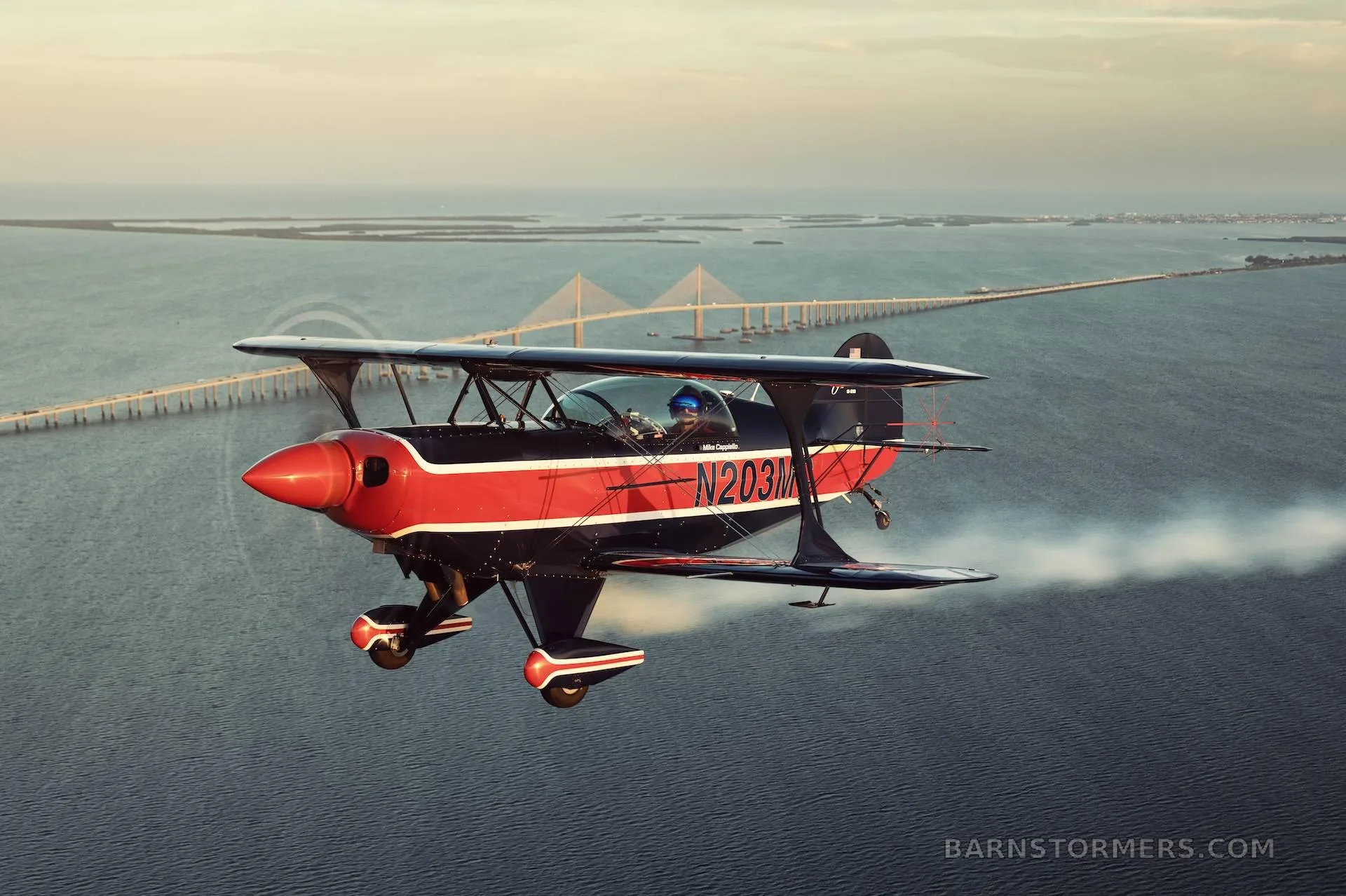 A dark blue and red biplane with registration N203MC flies over water, leaving a smoke trail, with a long bridge in the background.