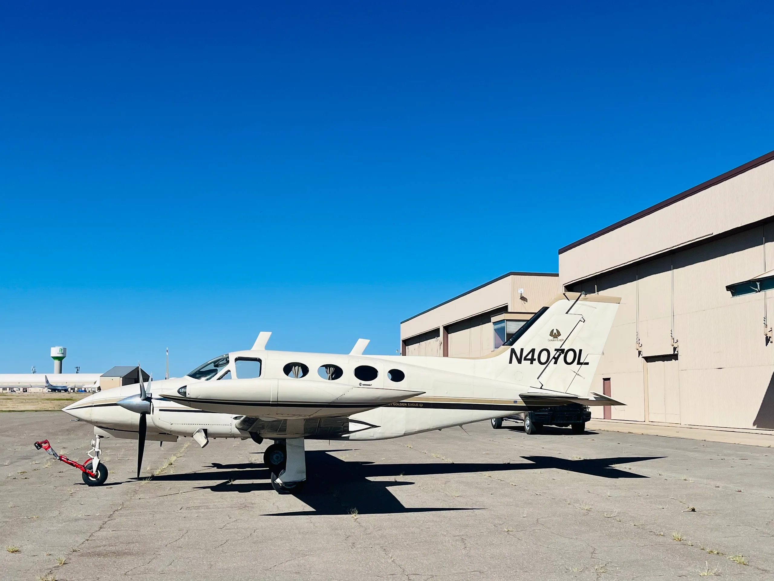 A white and gold Cessna 421 Golden Eagle (N4070L) parked on an airfield next to hangars under a clear blue sky.