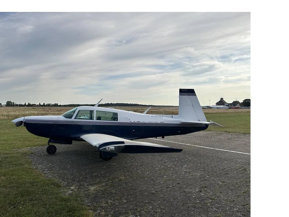 A blue and white single-engine aircraft parked on a tarmac with a grassy field and distant buildings under a cloudy sky.
