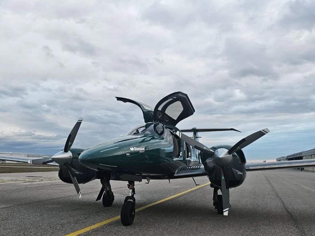 A dark green Diamond DA62 aircraft with its doors open is parked on a tarmac under a cloudy sky.