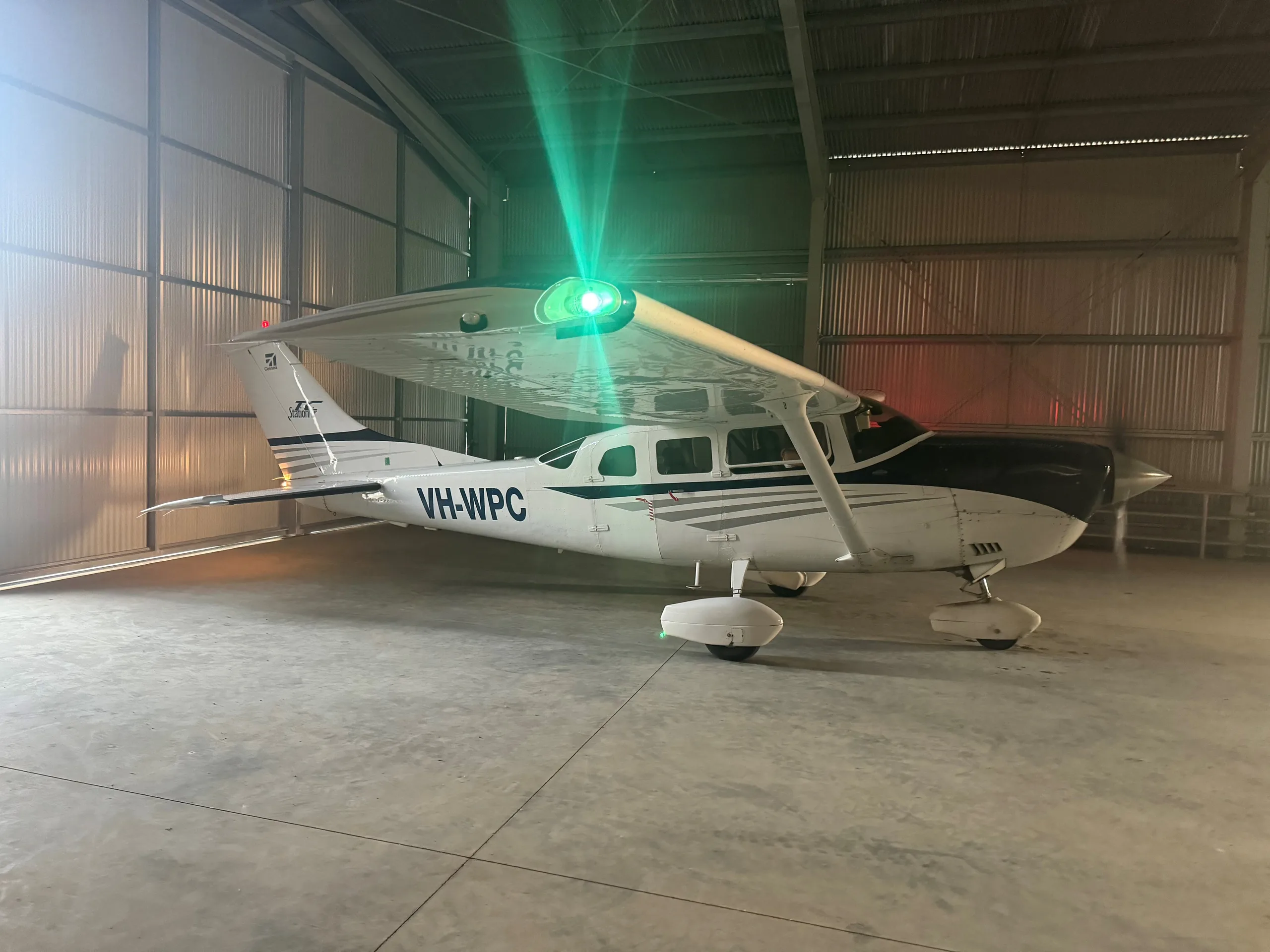 A white Cessna 210 with blue stripes, registration VH-WPC, parked inside a hangar with a green light beam shining from its wing.