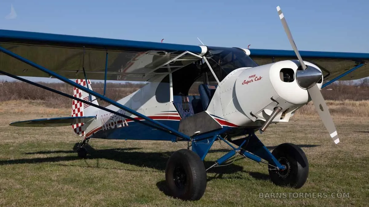 A white and blue Piper Super Cub aircraft with large tundra tires is parked on a grassy field under a clear sky.