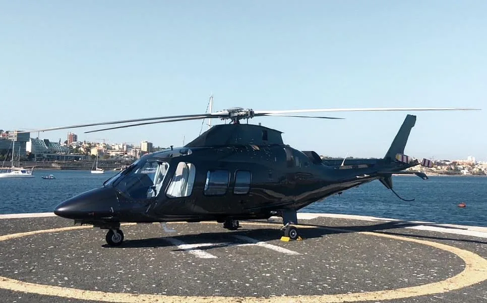 A dark grey helicopter parked on a circular helipad next to a body of water with boats and a city skyline in the background.