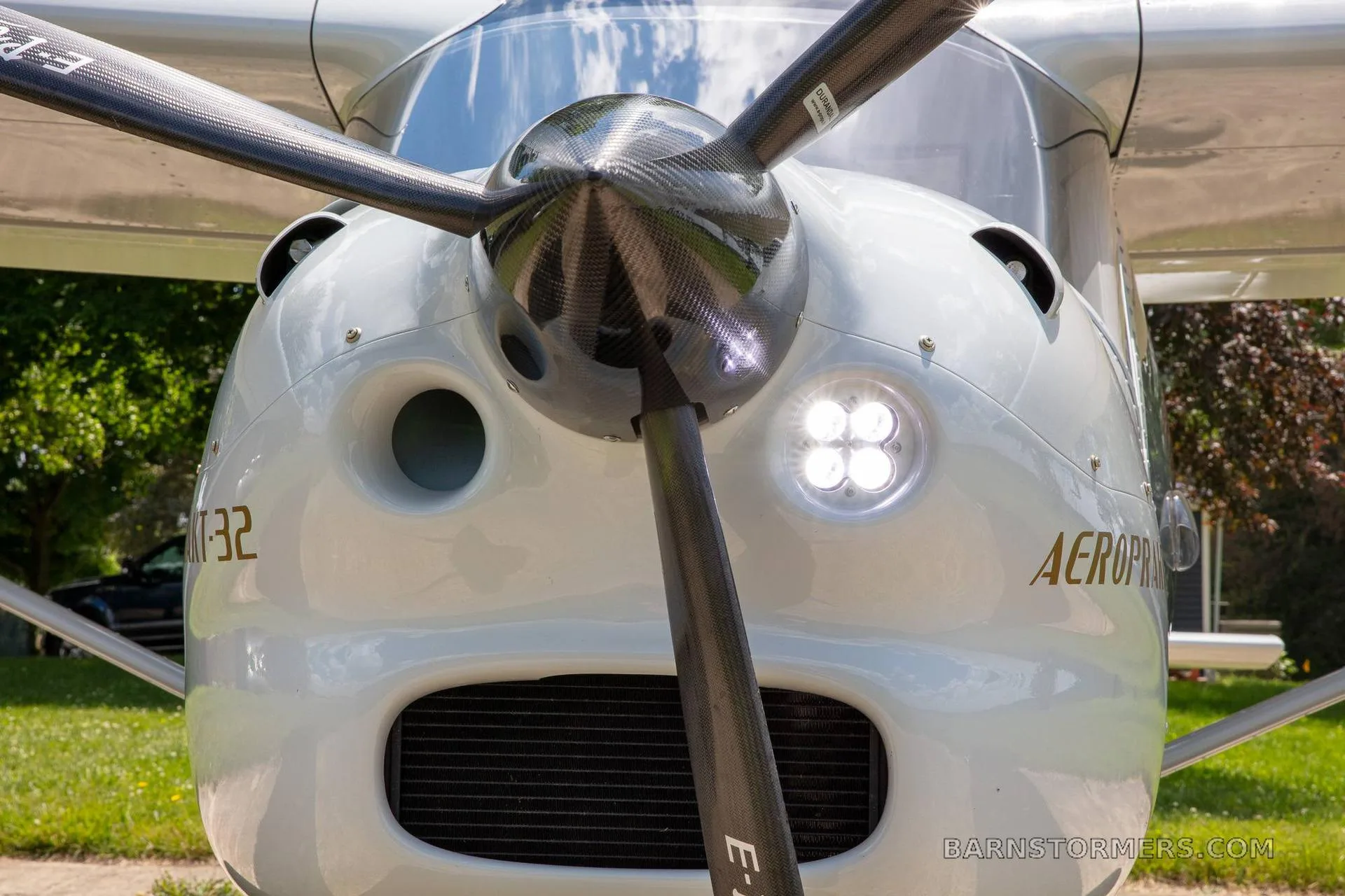 A head-on close-up view of the white Aeroprakt-32 aircraft's nose, featuring the propeller and bright landing lights.