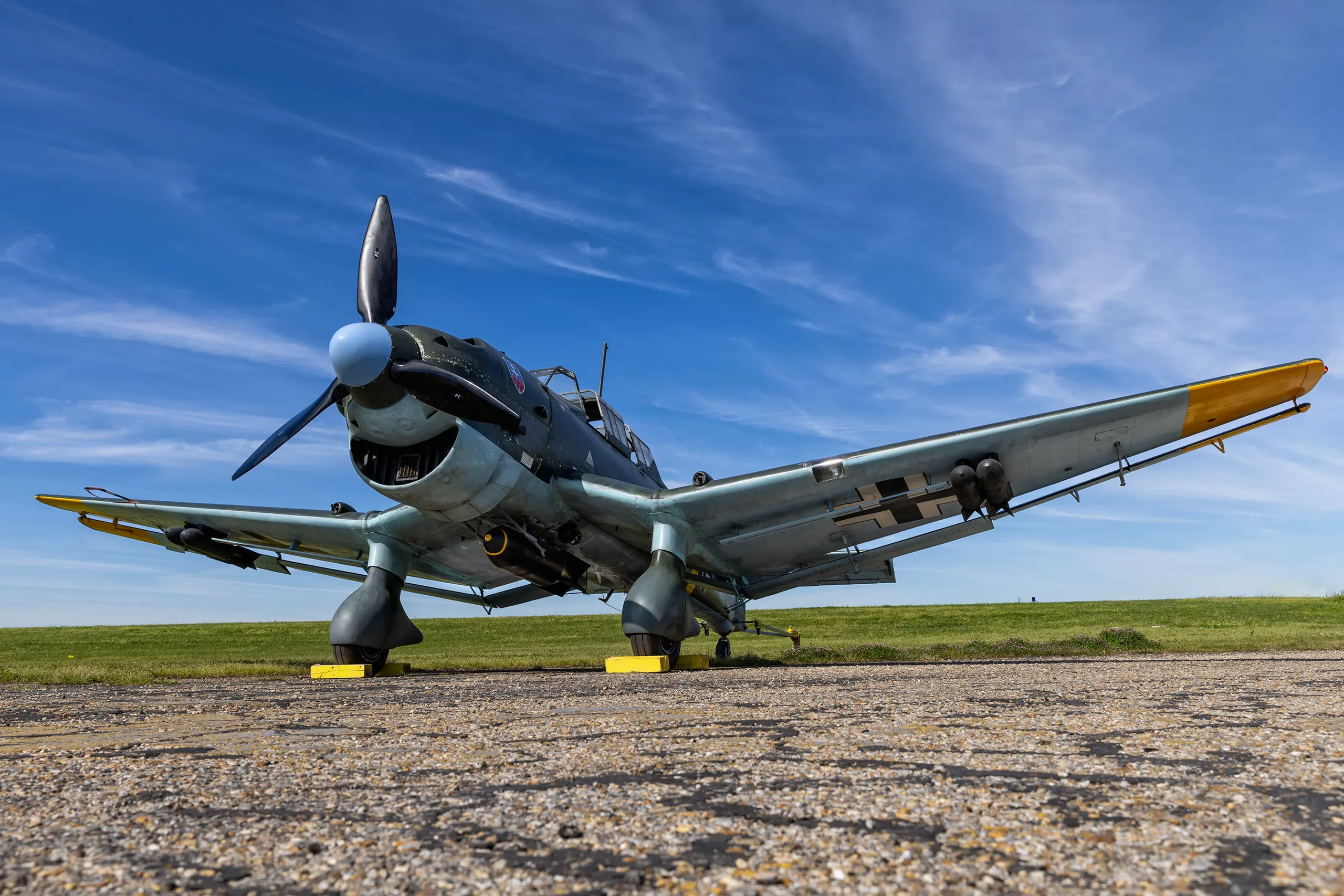 A low-angle view of a dark green and light blue Junkers Ju 87 Stuka dive bomber on an airfield under a blue sky.