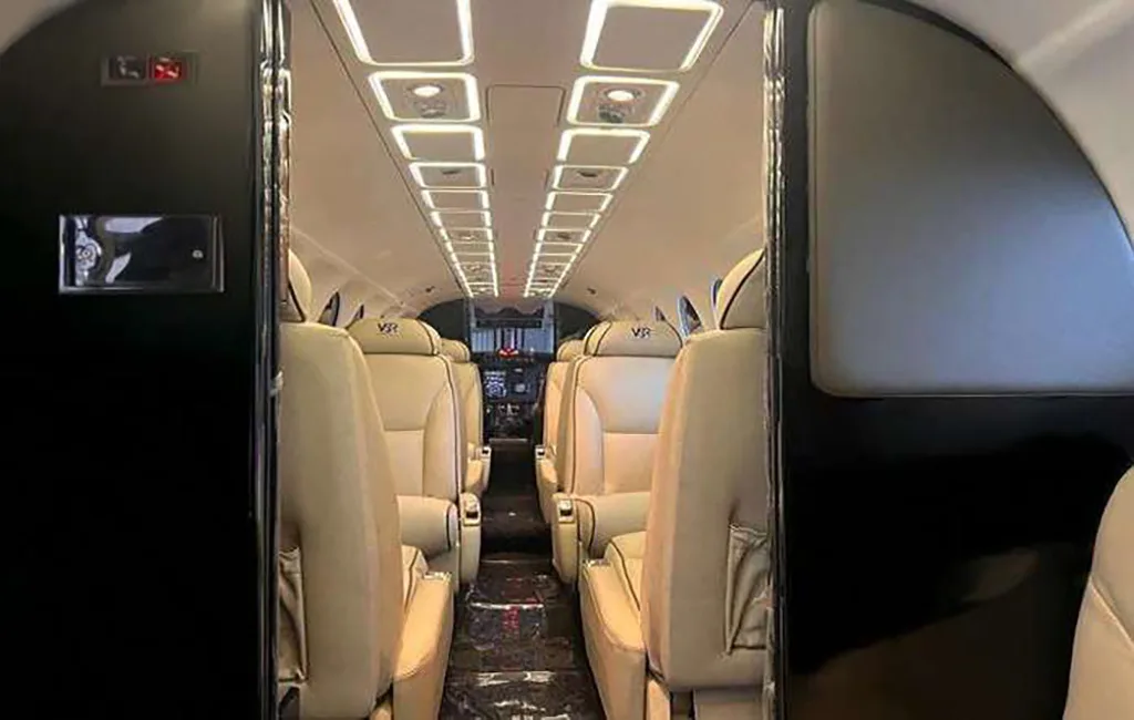 A view down the aisle of a private aircraft cabin with light beige seats and illuminated overhead panels.