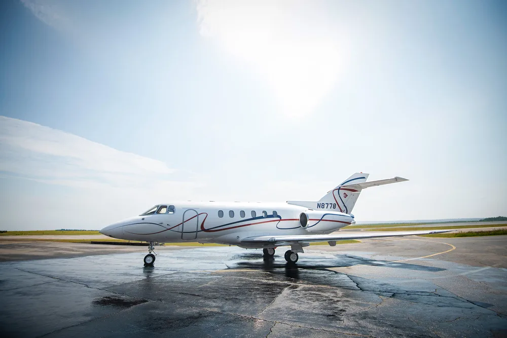 A white business jet with red and blue stripes (N8778) sits on a wet tarmac under a bright sky.