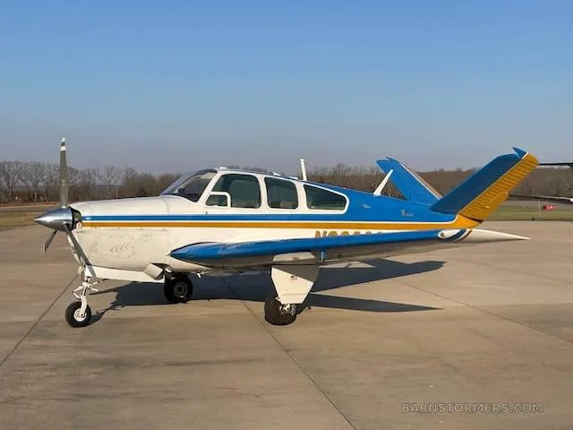 A white, blue, and gold Beechcraft Bonanza with tail number N8693Q is parked on an asphalt ramp under a clear blue sky.