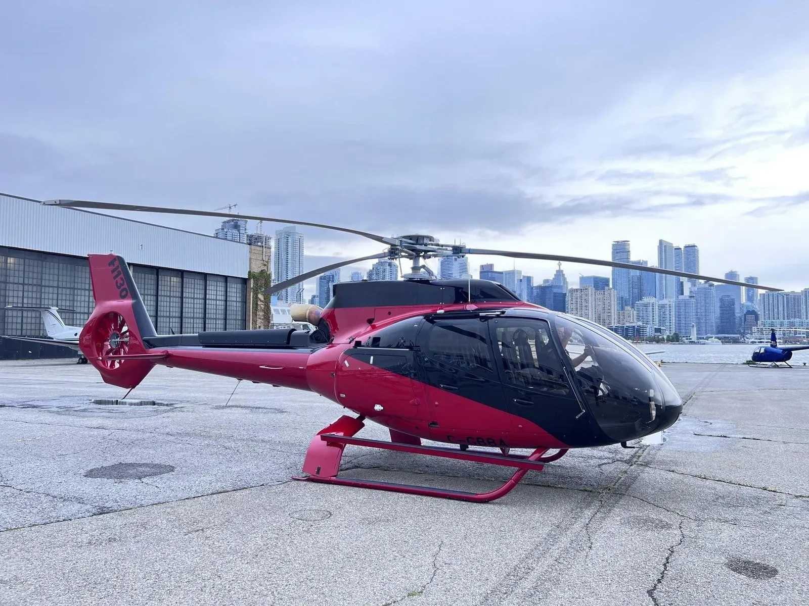 A red and black Airbus H130 helicopter parked on an airfield with a city skyline in the background.