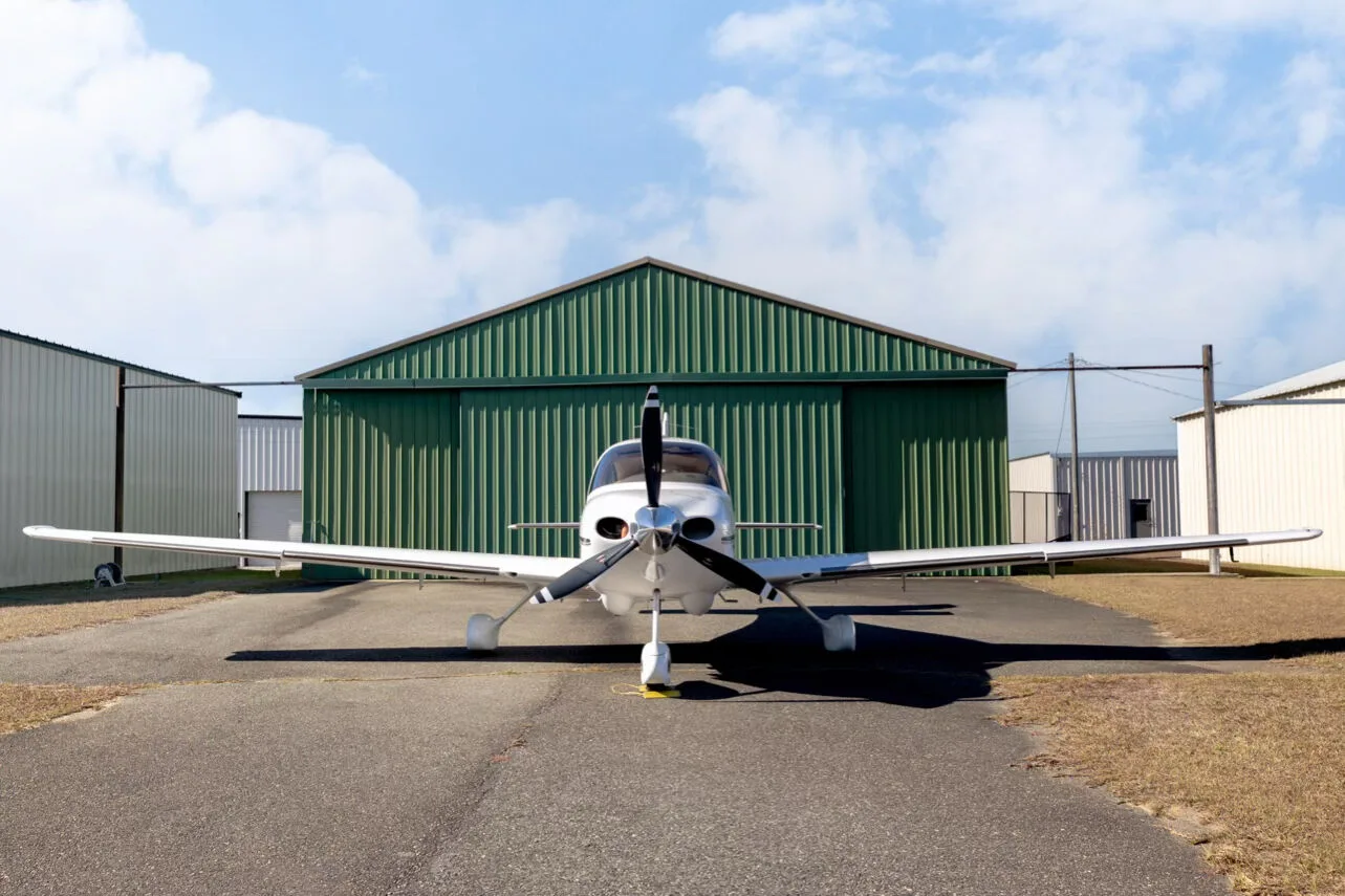 Front view of a white Cirrus SR22 aircraft with a three-blade propeller, parked in front of a green hangar.