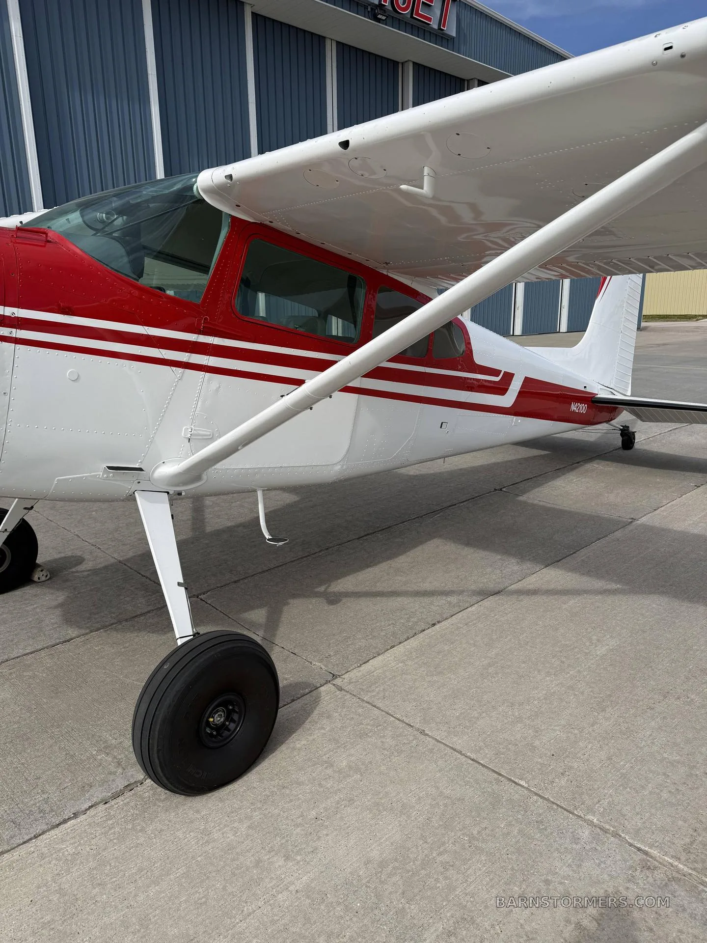 A close-up side view of the fuselage and wing of a white and red single-engine propeller aircraft (N42100) on an airfield.