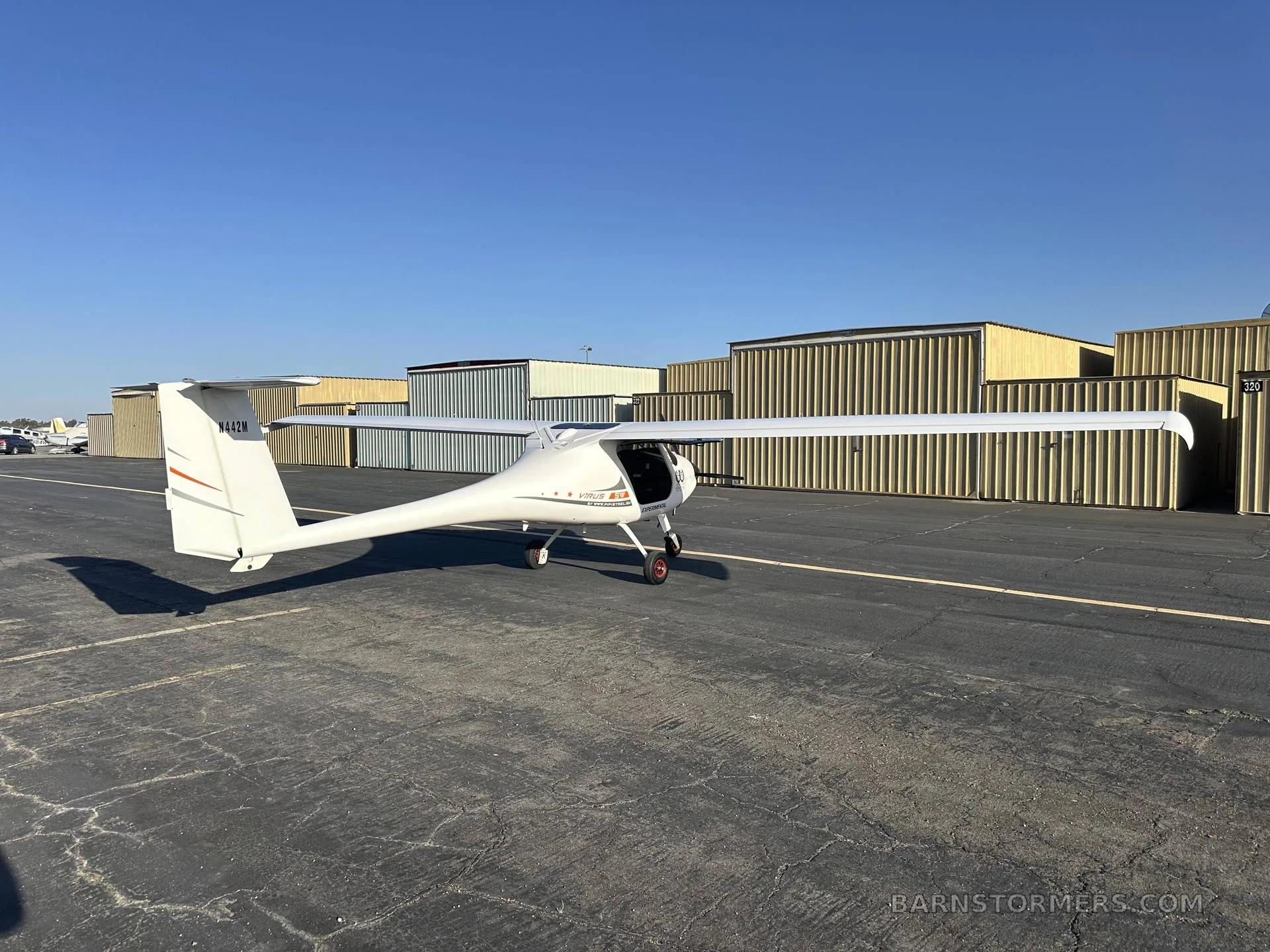 A white light sport aircraft, N442M, with orange stripes on the tail, viewed from the side on an asphalt tarmac with hangars in the background.