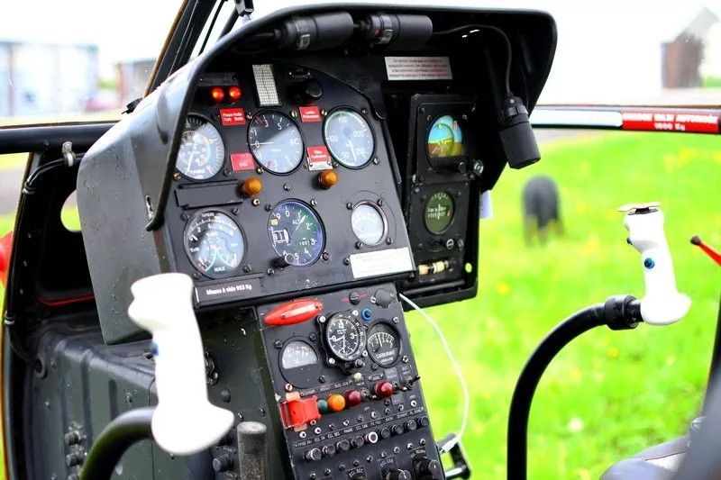 The instrument panel of a red helicopter, showing various gauges and controls, with a green field visible through the windshield.