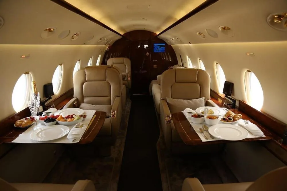 The luxurious interior cabin of a private jet with beige leather seats and tables set with food.