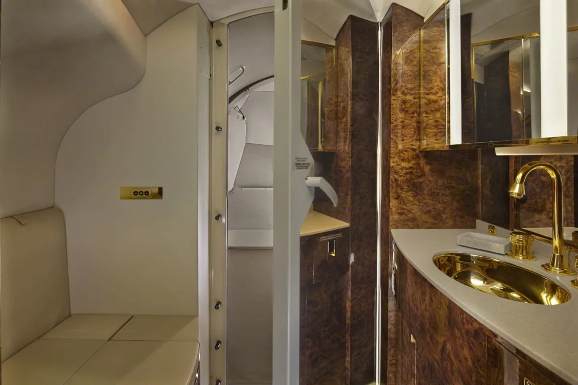A private jet lavatory with a gold sink and faucet, polished wood paneling, and a small seating area.