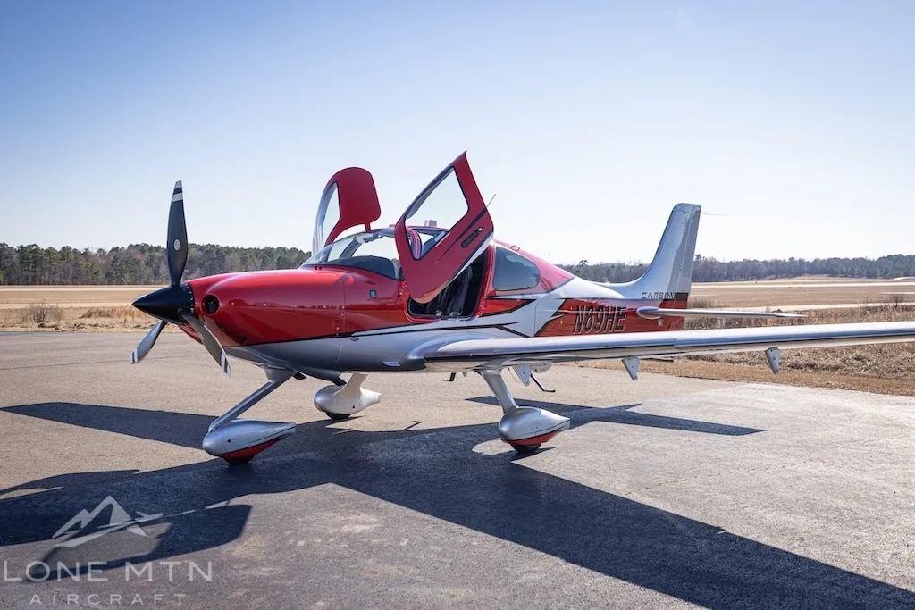 A red and silver Cirrus SR22T aircraft with open doors parked on an asphalt ramp.