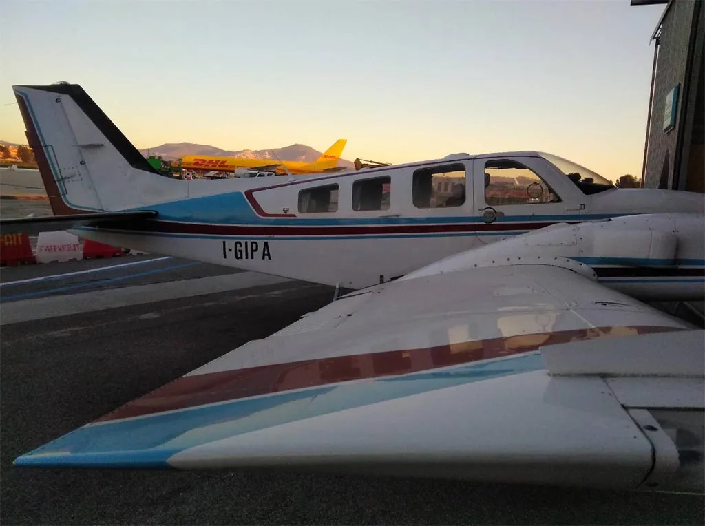 A white twin-engine aircraft with blue and red stripes, registration I-GIPA, parked on an airfield with a yellow DHL plane in the background at sunset.
