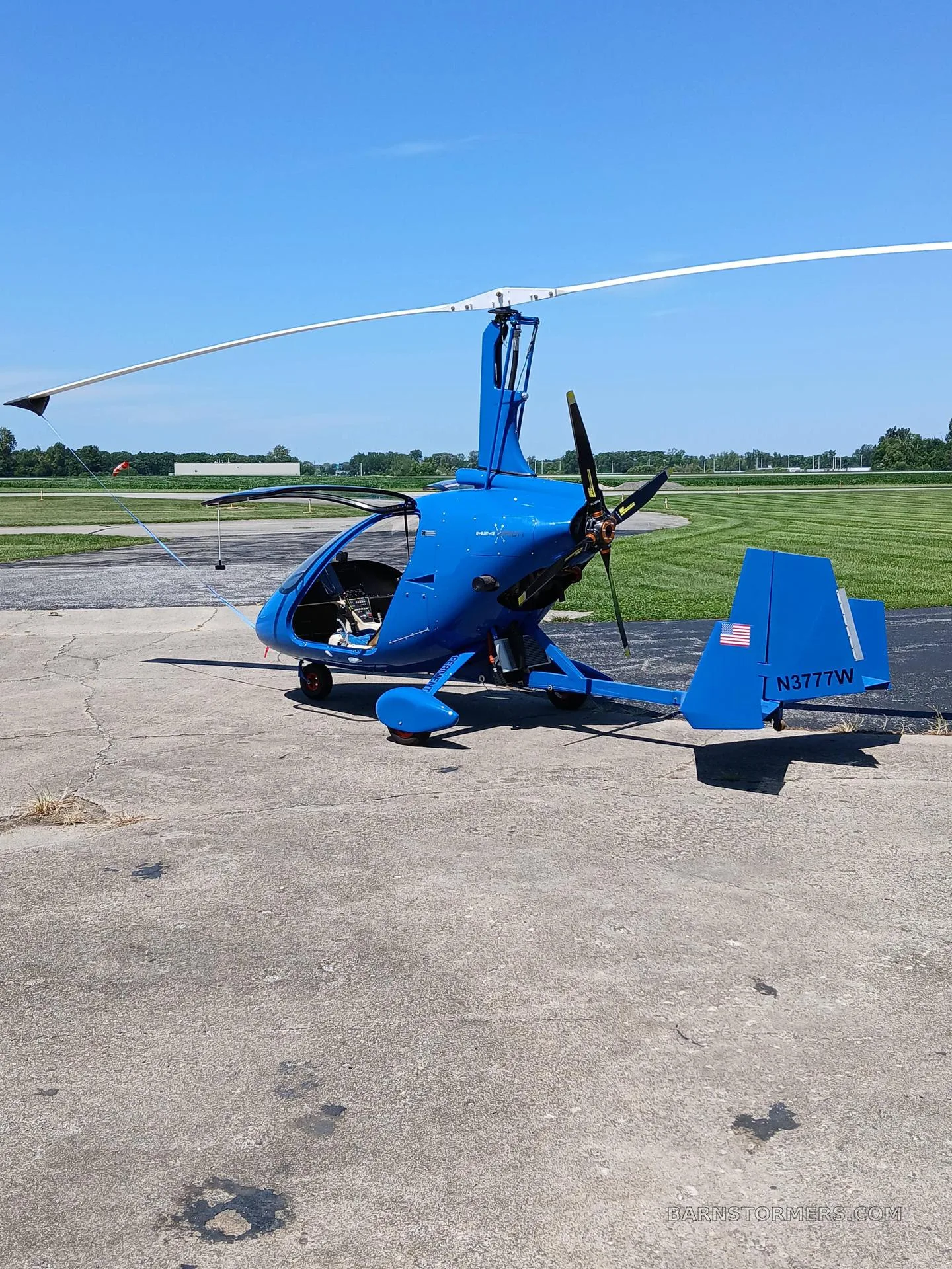 A blue gyrocopter with tail number N3777W parked on a concrete tarmac at an airfield on a sunny day.