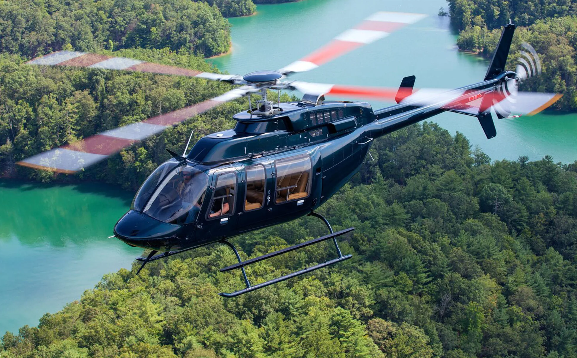 A dark green helicopter with red and white main rotor blades flies over a lush green forest and a turquoise lake.
