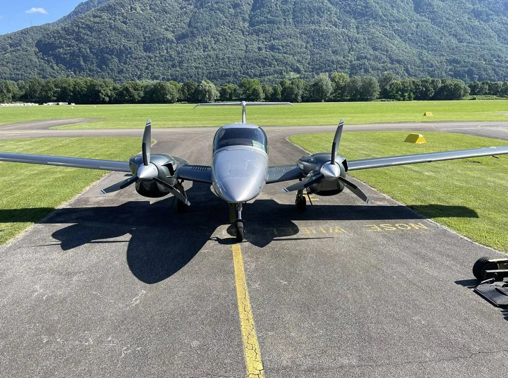 A silver Diamond DA42 aircraft parked head-on on an airfield runway with green fields and mountains in the background.