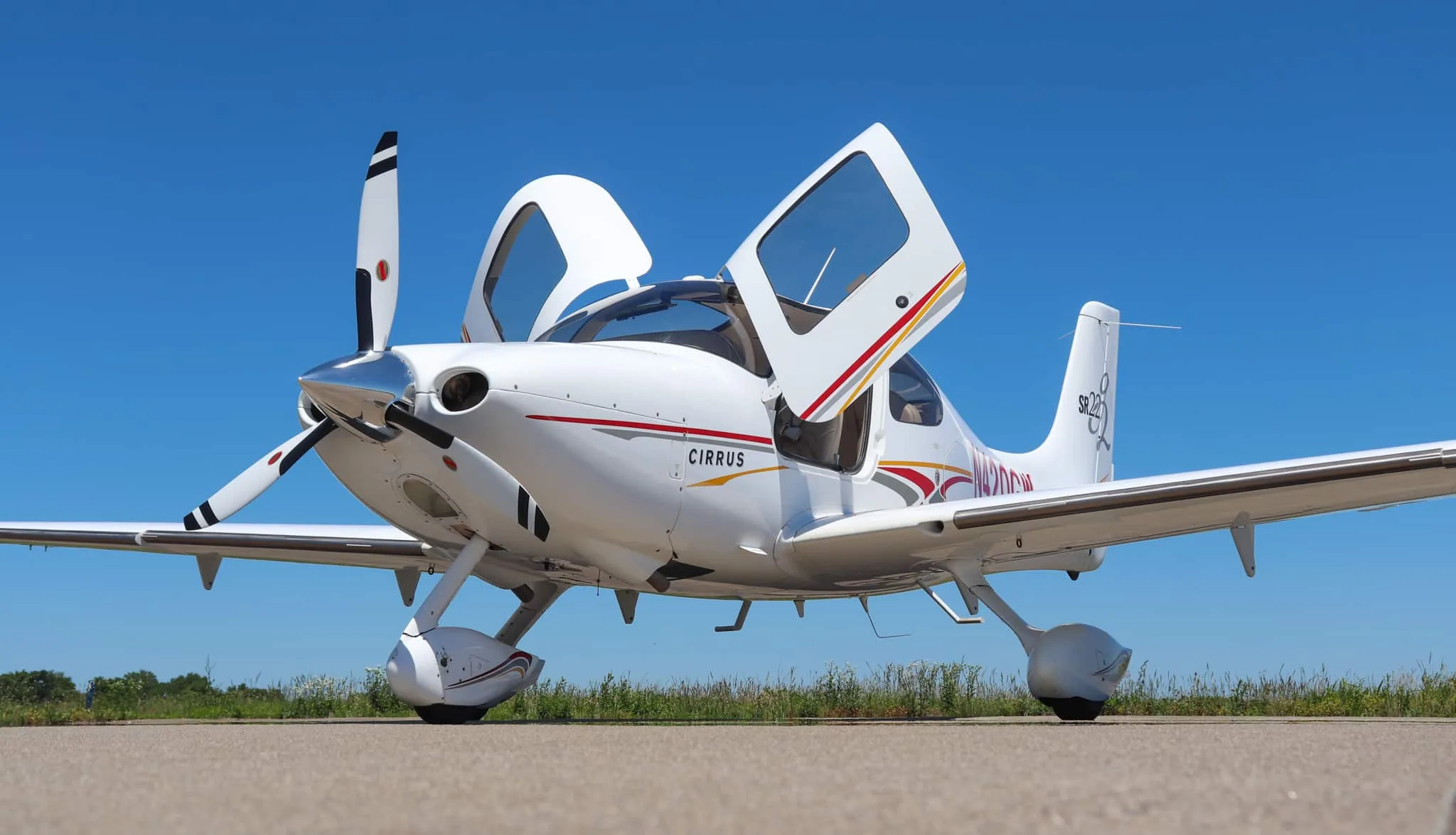 Front-left view of a white Cirrus SR22 aircraft with open gull-wing doors on a tarmac.