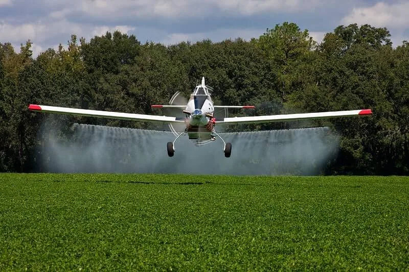 A white and red agricultural aircraft sprays a green field from a low altitude, with trees in the background.
