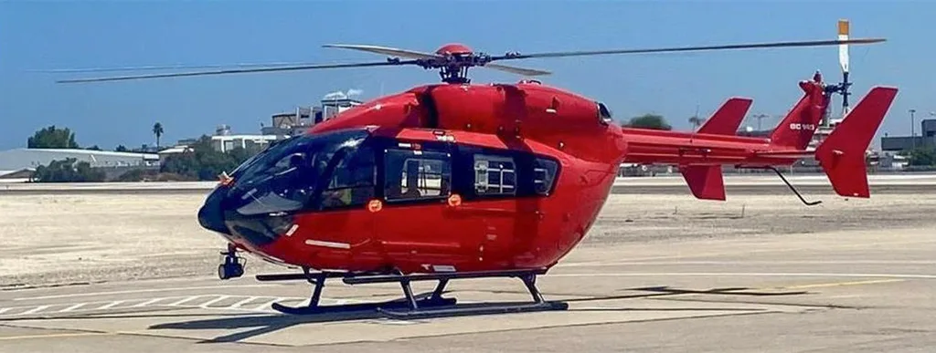 A red Airbus H135 helicopter with black skids and tinted windows parked on a sunny airfield.