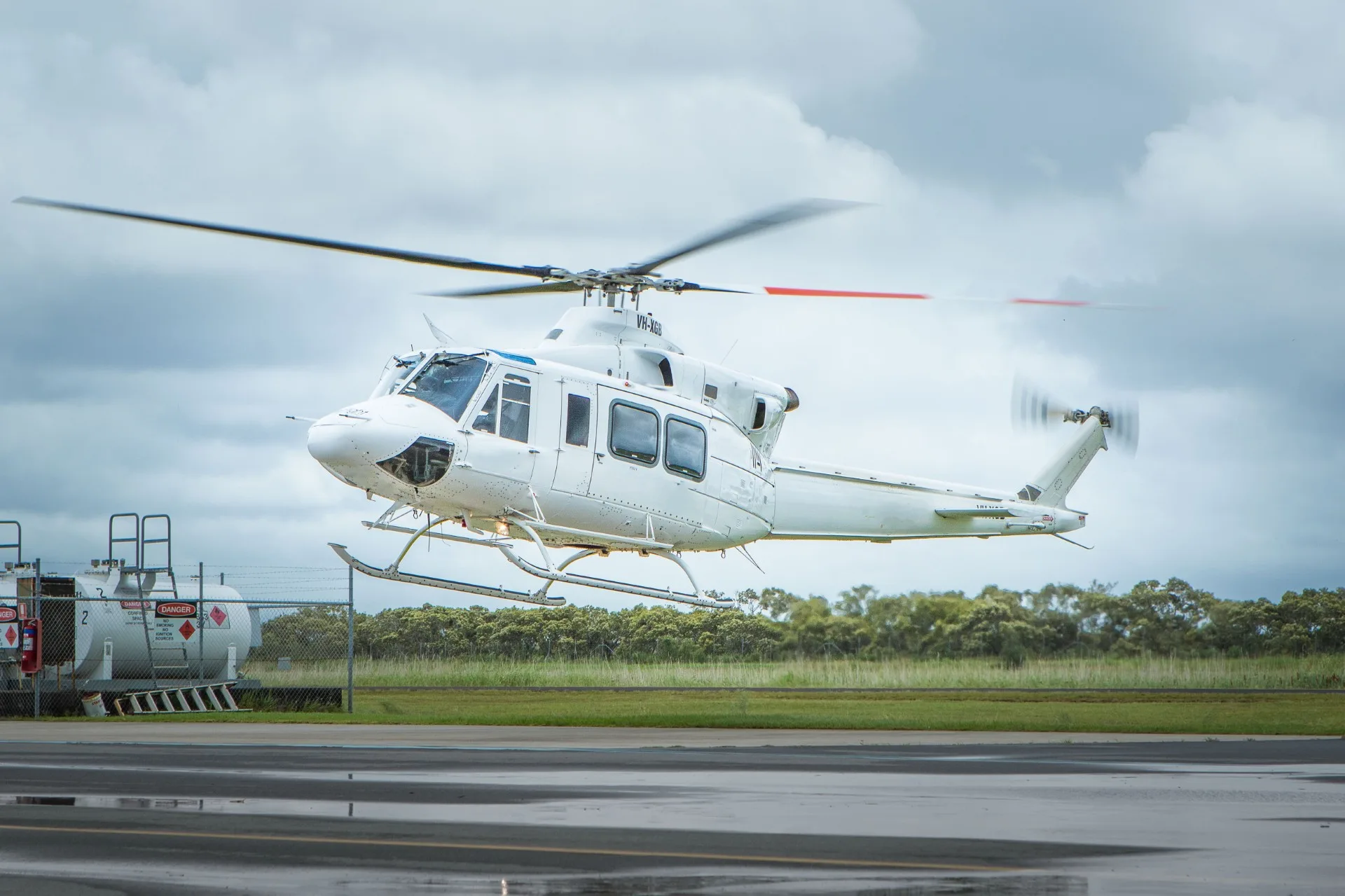A white twin-engine helicopter, possibly a Bell 412, hovers just above a wet tarmac with fuel tanks and trees in the background under a cloudy sky.