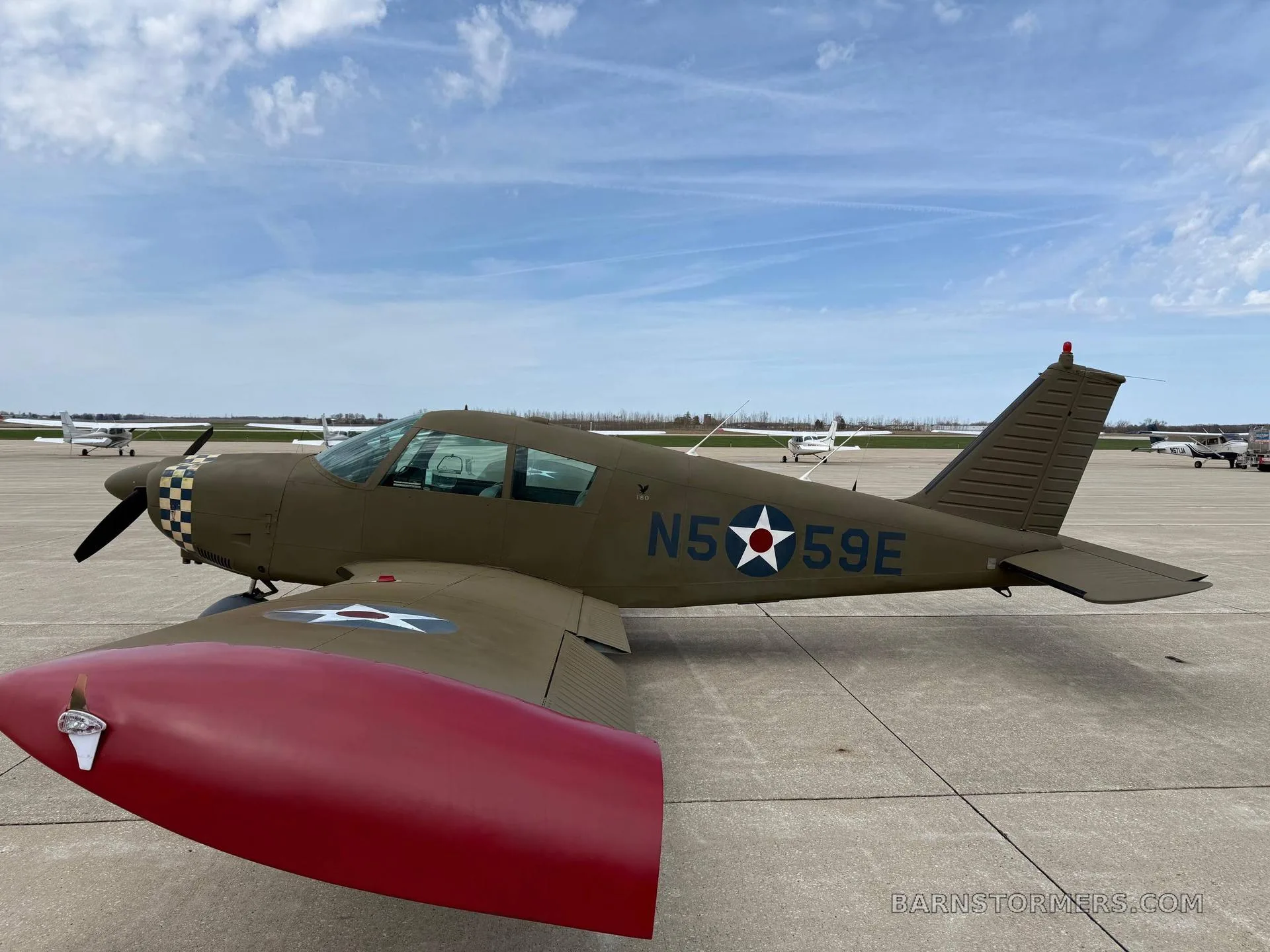 A green single-engine propeller aircraft with a red wingtip and checkered nose, parked on an airfield under a partly cloudy sky.