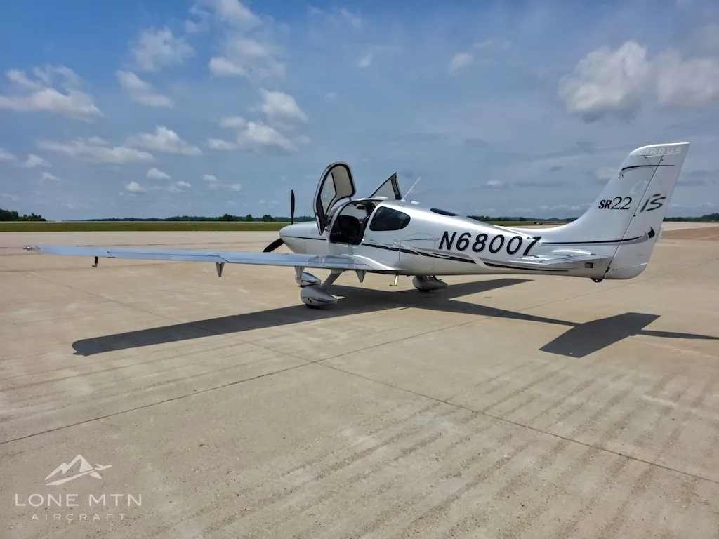 A silver Cirrus SR22 aircraft (N68007) with its gull-wing doors open, viewed from the rear-left on an airfield.