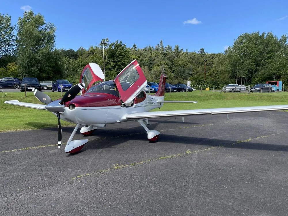 A red and white Cirrus SR22 aircraft with its gull-wing doors open is parked on an airfield.