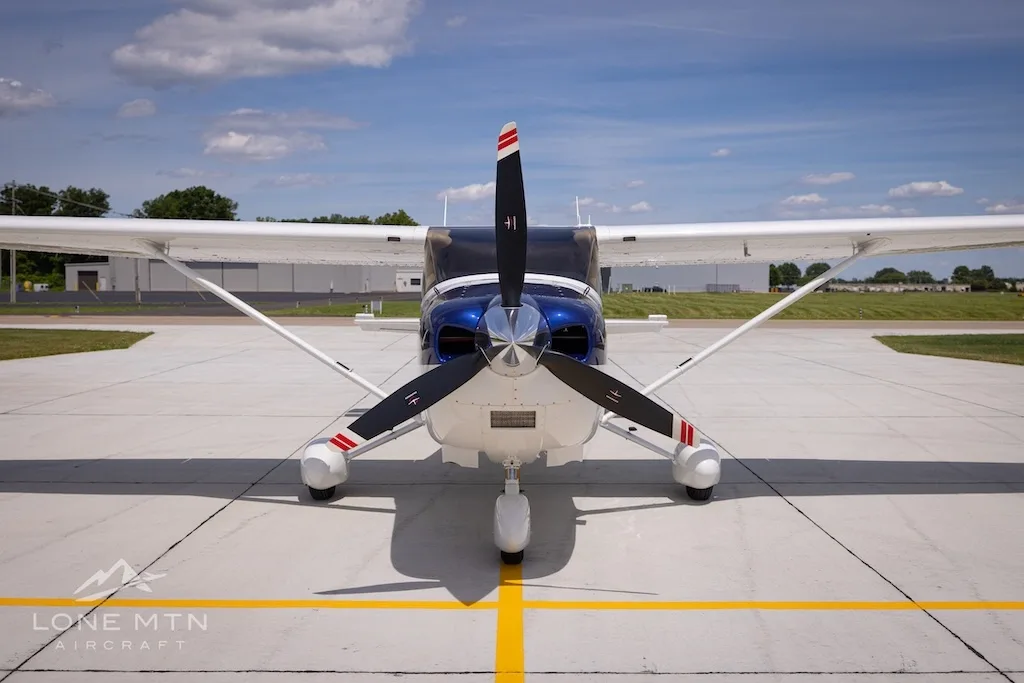 A head-on view of a white and blue Cessna 182 Skylane (N27GH) with its propeller facing forward on an airport tarmac.
