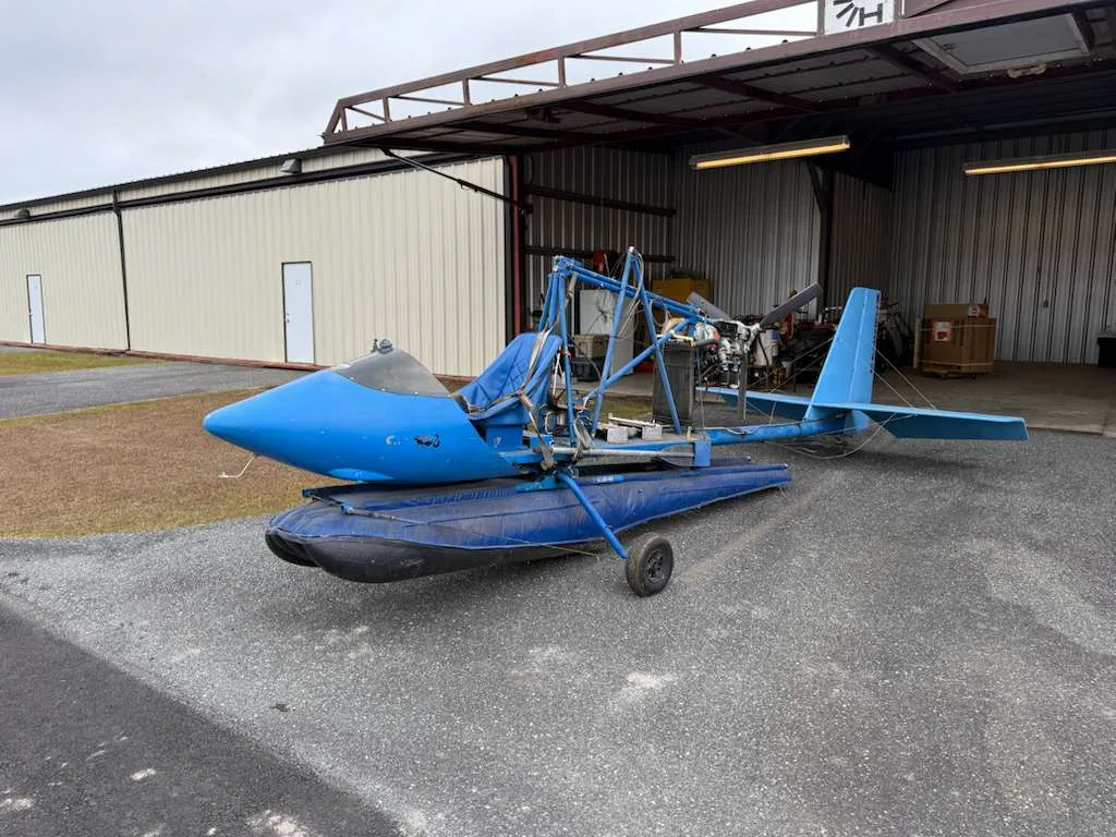 A blue ultralight aircraft on floats, with its engine exposed, parked on gravel outside a hangar.