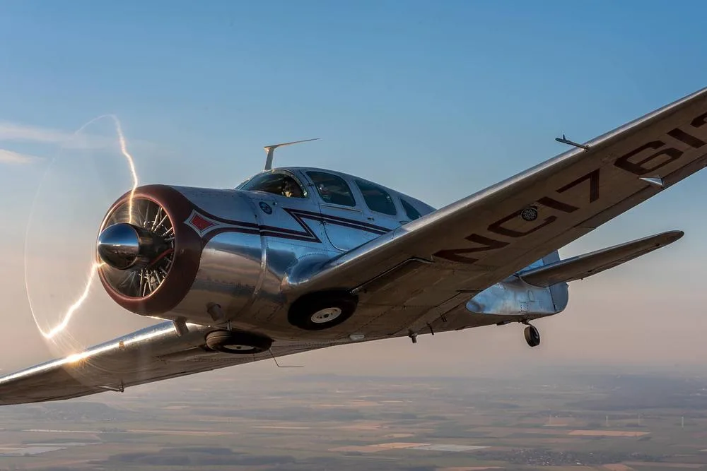 A silver vintage Beechcraft Staggerwing aircraft with a red nose cone in flight, showing registration N17613 on the wing.