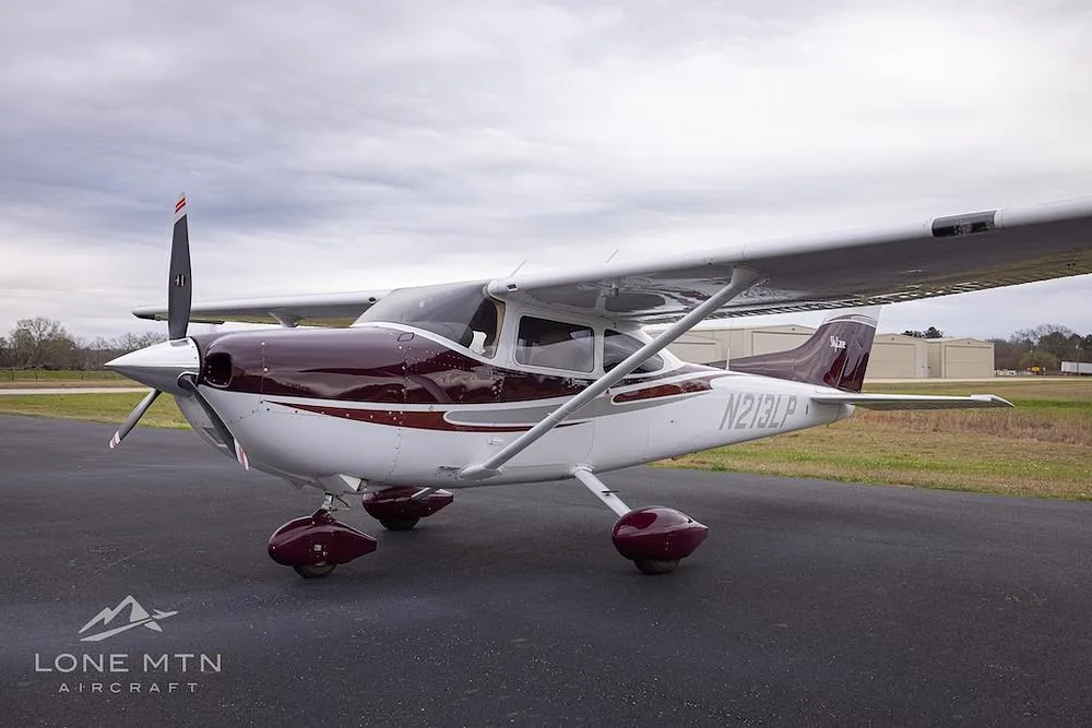 A Cessna 182 Skylane with a maroon and white paint scheme, parked on an asphalt ramp under a cloudy sky, viewed from the front-left.