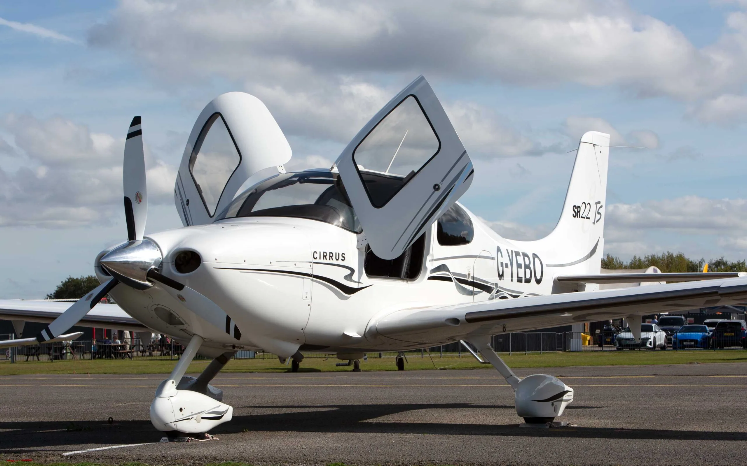 A white Cirrus SR22 TSi aircraft with black and grey stripes, registration G-YEBO, parked on an airfield with its gull-wing doors open.