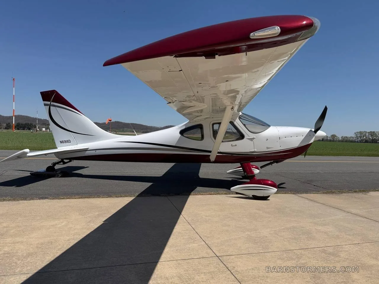 A white and maroon high-wing single-engine aircraft (N686D) is parked on an airfield with a runway and hills in the background.