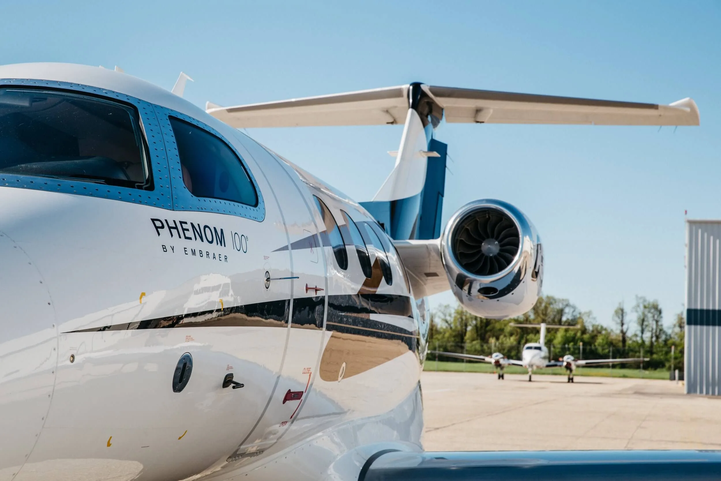 A close-up view of the rear fuselage and engine of a white Embraer Phenom 100 jet on an airfield.