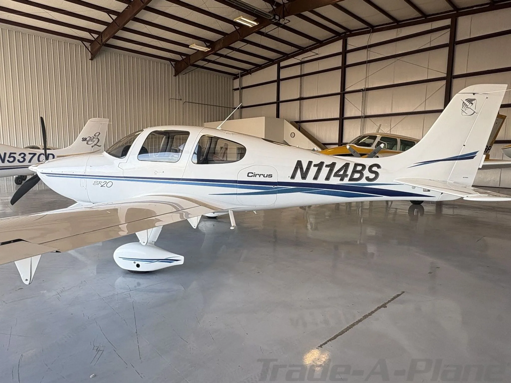 A white Cirrus SR20 aircraft with blue stripes, registration N114BS, parked in a hangar.