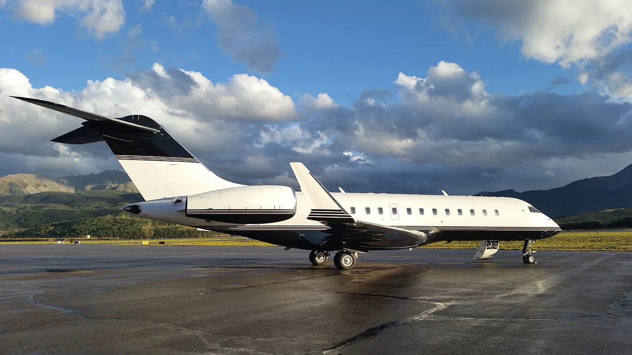 A white business jet with black tail and accents is parked on a wet tarmac with mountains and a cloudy sky in the background.