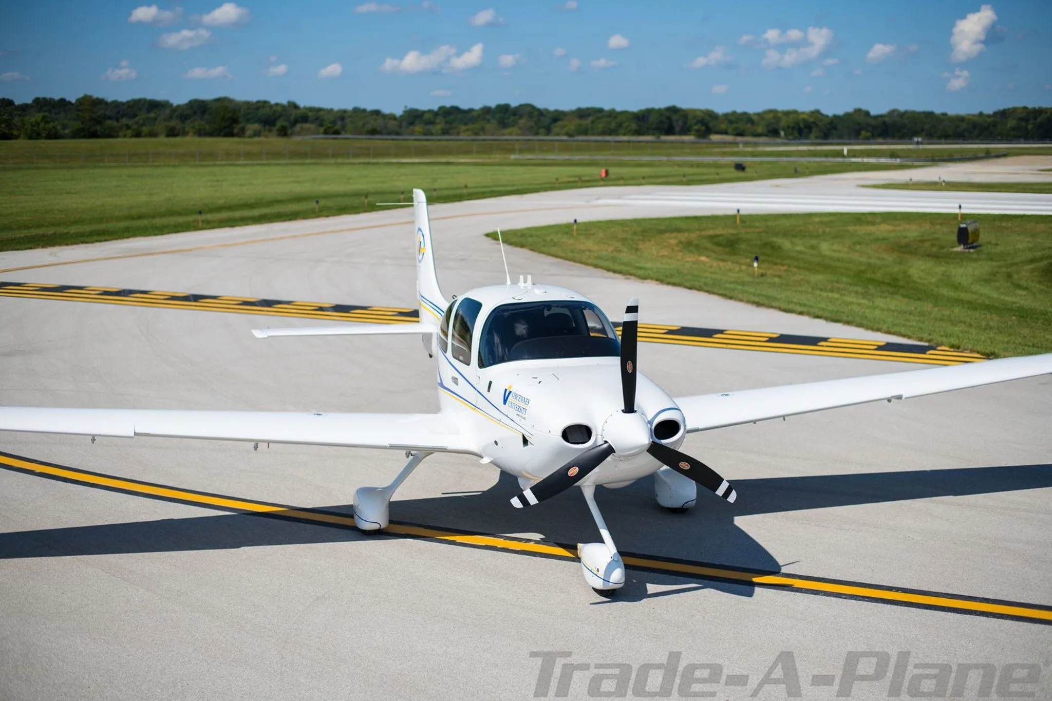 A white single-engine propeller aircraft, N42VU, with blue and yellow stripes, parked on an airfield with green fields in the background.