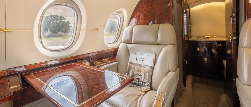 A close-up view of the business jet's cabin, showing a beige leather seat, a polished wood table, and the entrance to the lavatory.
