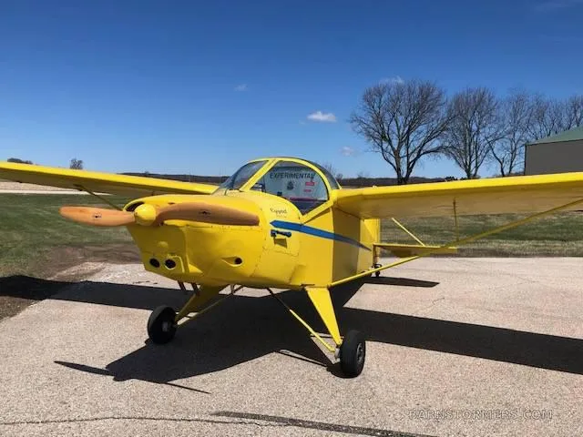 A bright yellow single-engine experimental aircraft with a wooden propeller, viewed from the front-left, parked on an asphalt surface.