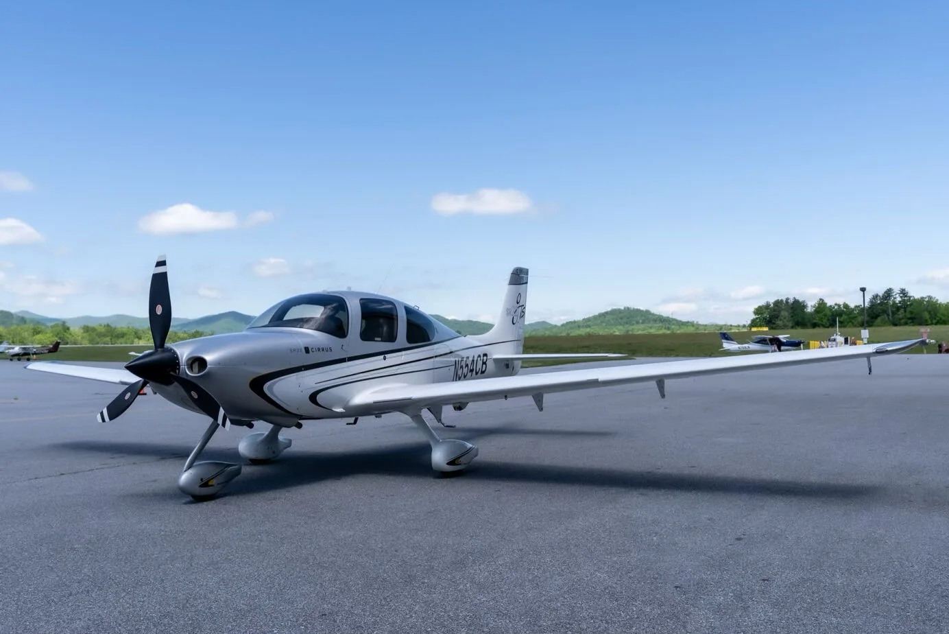 A silver and white Cirrus SR22T aircraft with registration N554CB parked on an airport tarmac with mountains in the background.